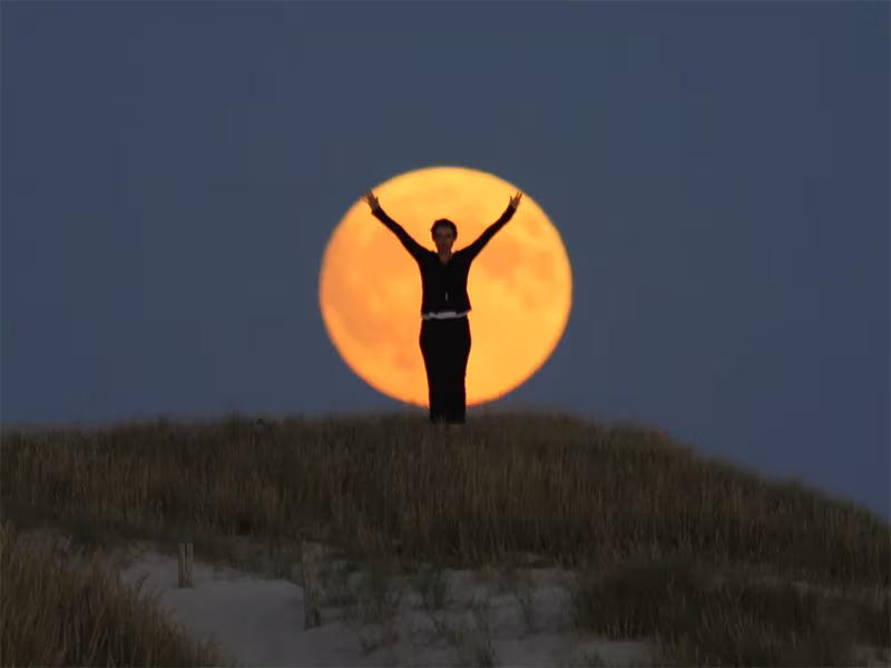Silueta de una persona con los brazos en alto recortada contra una enorme luna llena naranja