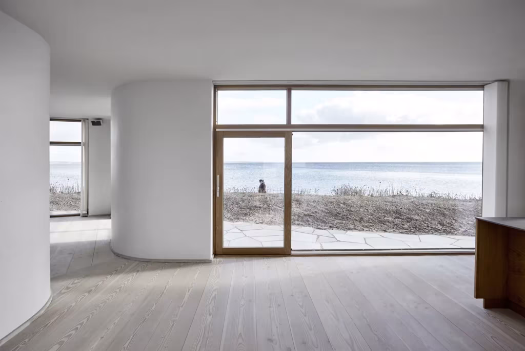 Sala diáfana de la Villa Platan con muro curvo blanco, suelo de madera clara y cristalera hacia el mar
