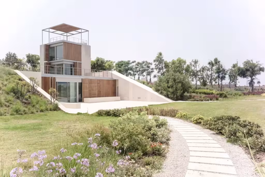 Vivienda de The Courtowers con su torre sobre una rampa verde y un sendero de losas hacia el mar