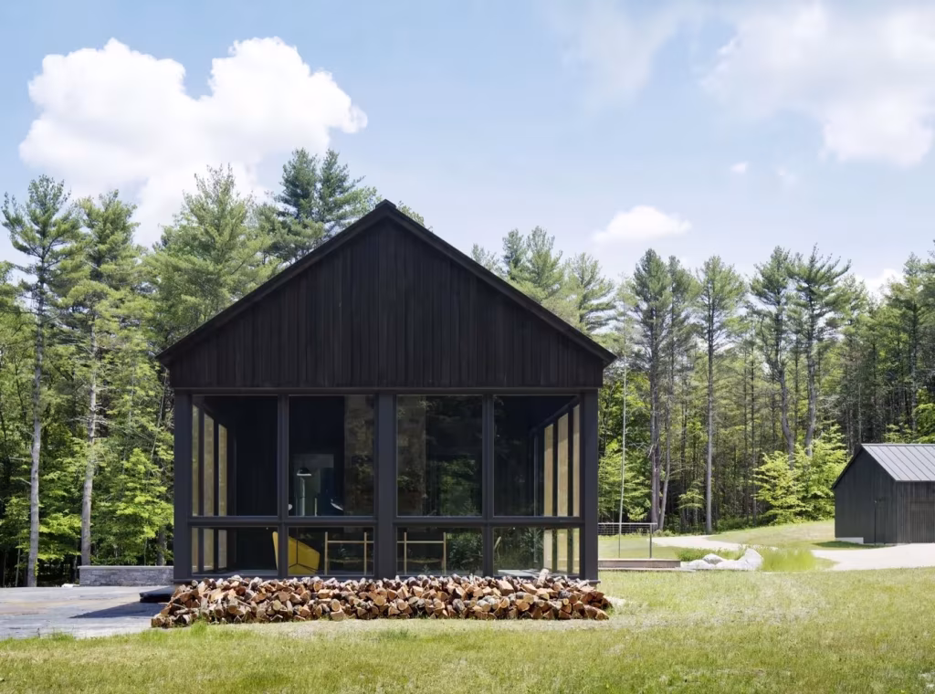 Casa de madera oscura a dos aguas con porche de cristal frente al bosque y un cobertizo al fondo