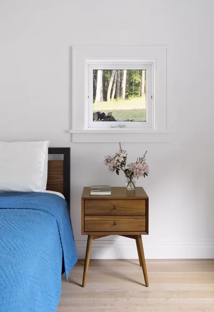 Dormitorio blanco con cama de colcha azul, mesilla de madera y una ventana pequeña al arbolado