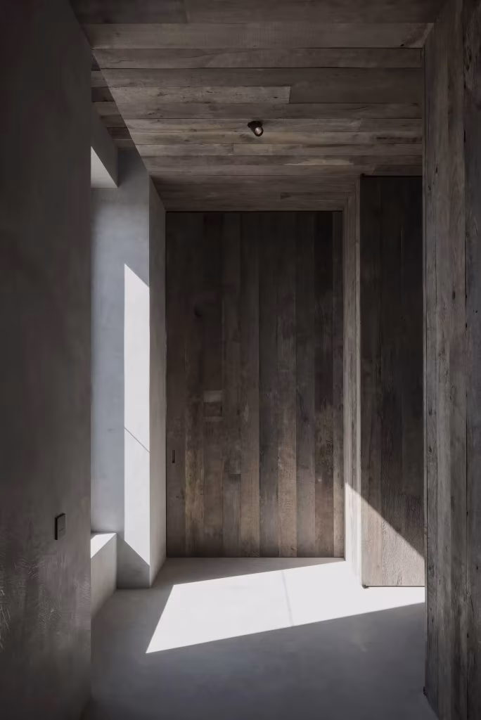 Corredor del C Penthouse con puerta de madera envejecida y luz oblicua sobre el suelo de microcemento