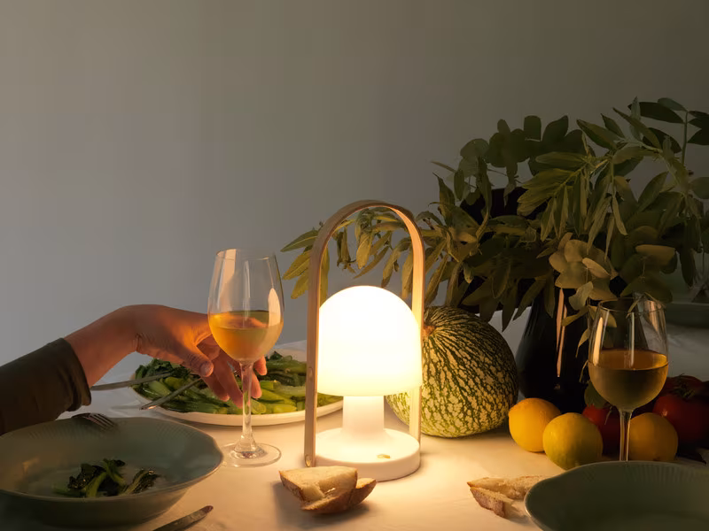 Lámpara Follow Me encendida sobre una mesa de cena con copas de vino, frutas y plantas