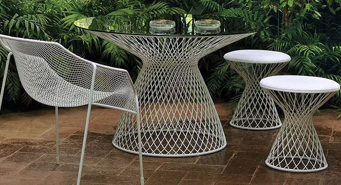 Mesa de exterior Heaven de malla metálica blanca con taburetes y silla ante vegetación tropical