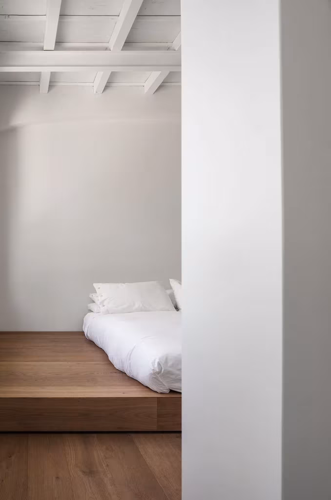 Dormitorio de la Casa AG con cama baja sobre plataforma de madera tras un muro blanco