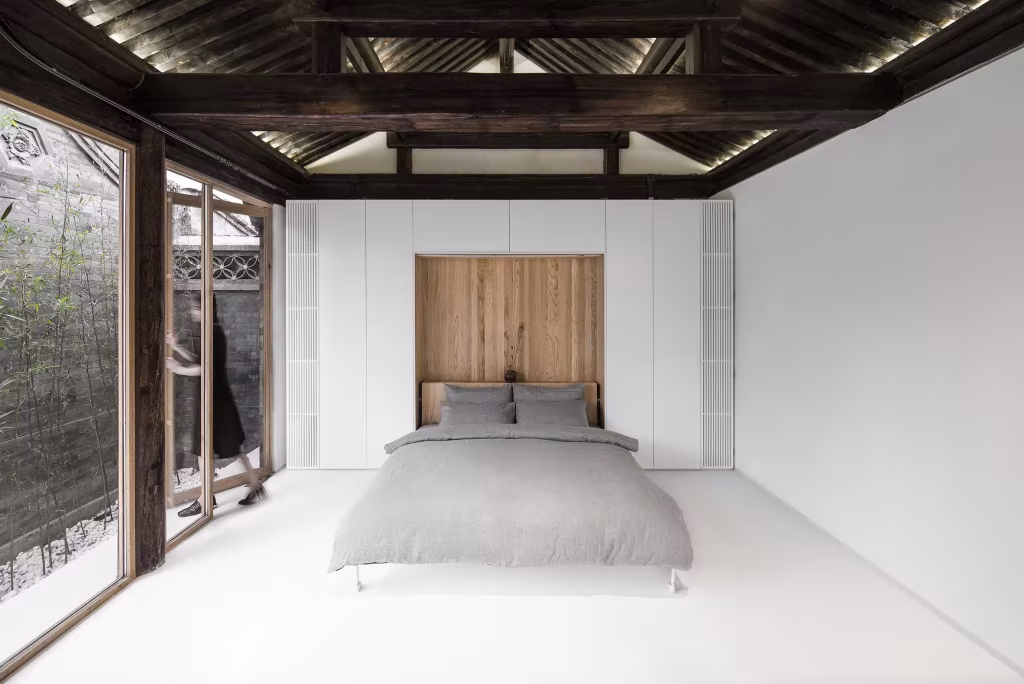 Dormitorio blanco bajo la estructura de madera original del siheyuan, con cama gris y cabecero de madera