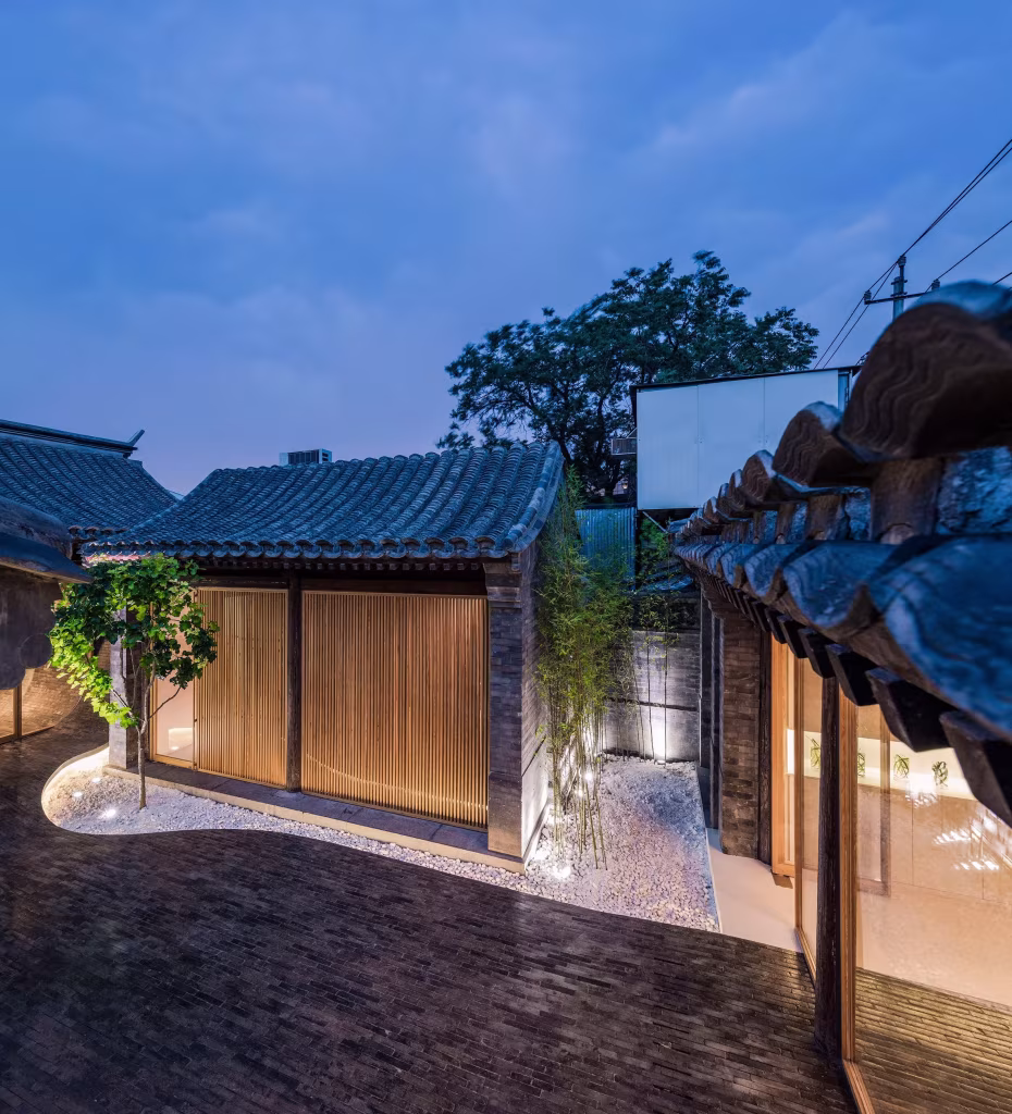 Patio del siheyuan al anochecer con tejados de teja gris, frentes de madera iluminados y un árbol