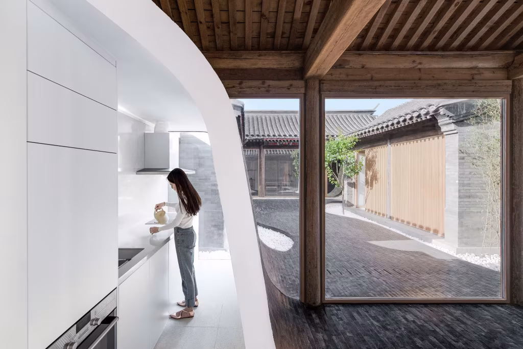 Cocina blanca de pared curva abierta al patio de ladrillo de la Twisting Courtyard House, con una persona cocinando
