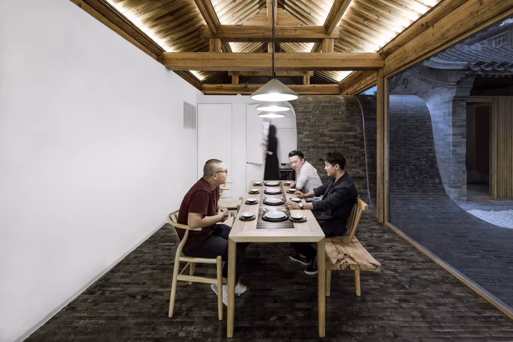 Comedor de la Twisting Courtyard House con mesa larga de madera, comensales y techo de vigas iluminado