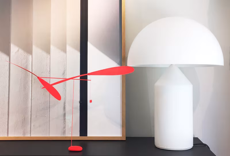 Móvil rojo de dos hélices junto a una lámpara Atollo blanca de pantalla en cúpula, sobre un aparador negro