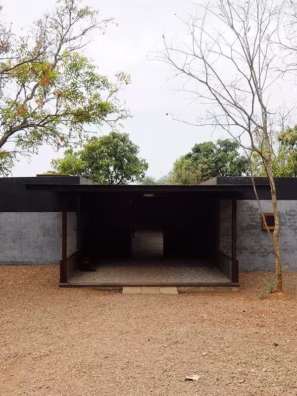 Pabellón de cubierta plana sobre pilares ante un muro gris, en un solar de tierra de Studio Mumbai