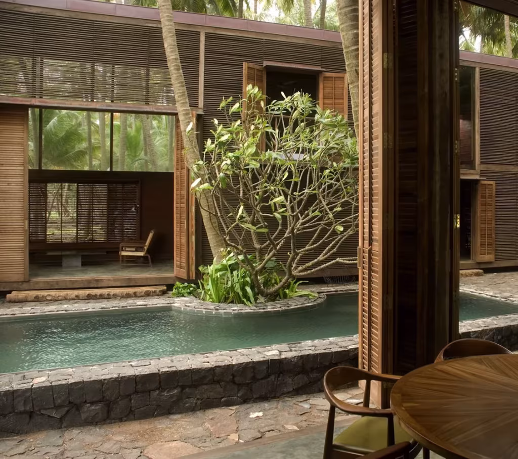 Patio con estanque, un árbol y fachada de madera entre pilares en la Palmyra House de Studio Mumbai
