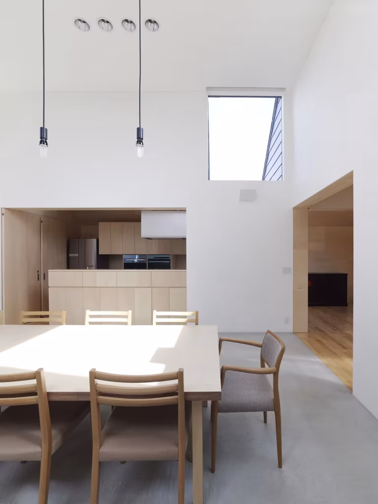 Comedor de doble altura con mesa clara, cocina de madera al fondo y luz cenital en la Casa Yagiyama