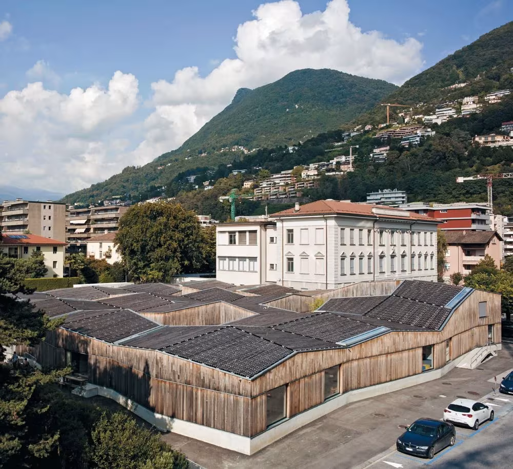 Vista aérea de las cubiertas quebradas del Kindergarten Cassarate de Lugano frente a un edificio histórico