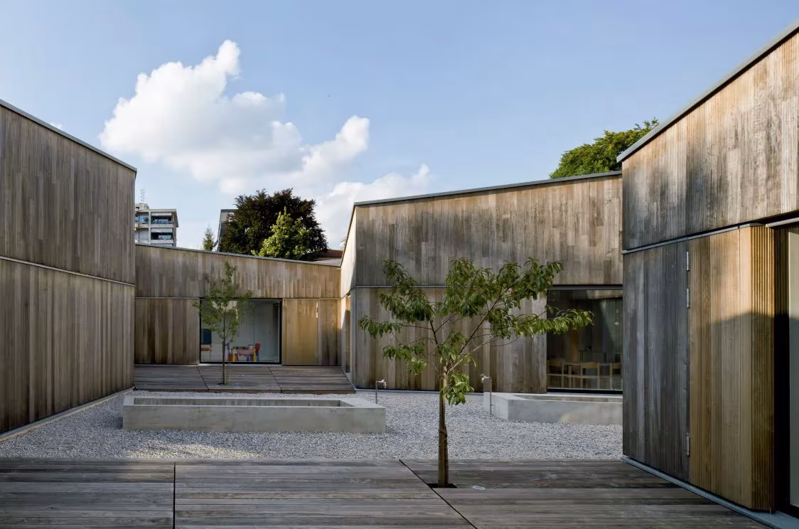 Patio de grava con estanque y árbol entre volúmenes de madera del Kindergarten Cassarate, en Lugano