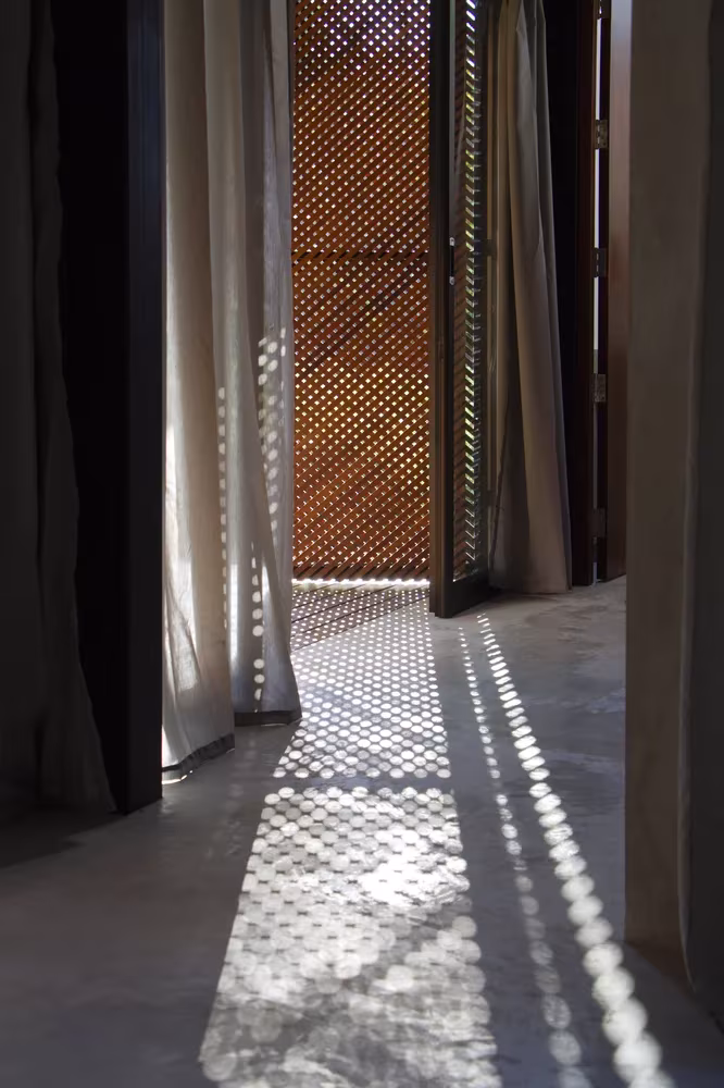 Pasillo con cortinas y panel de madera perforado que dibuja sombras sobre el suelo en la Phatthalung House