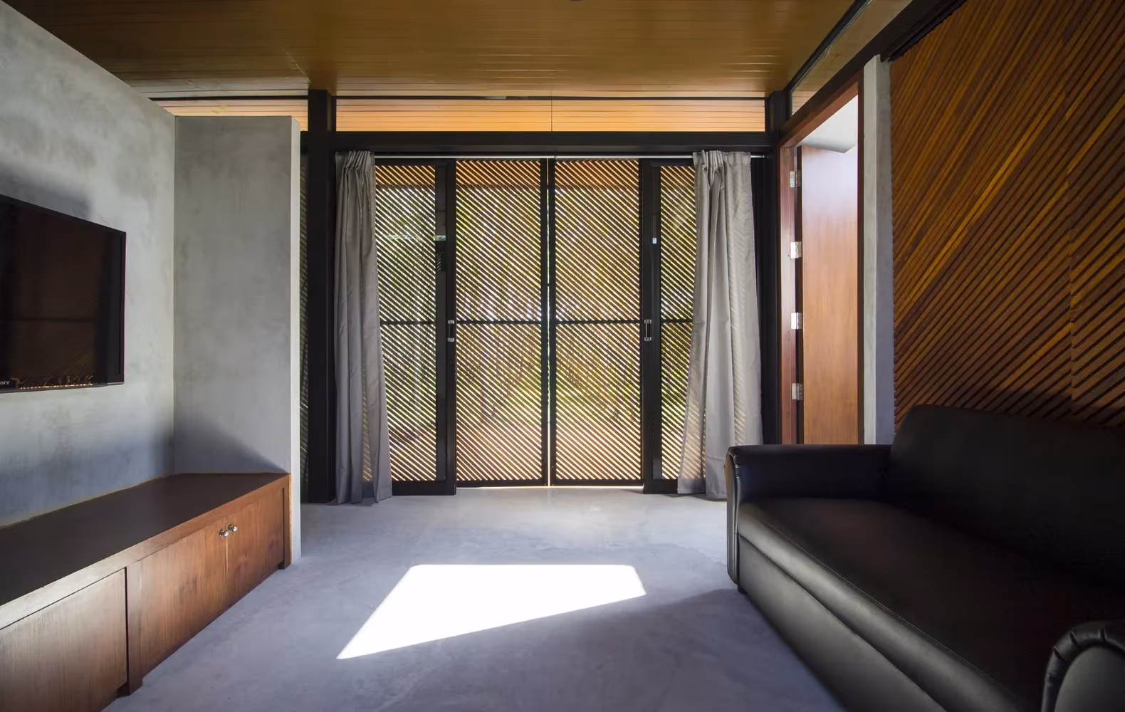 Sala con suelo de cemento pulido, mueble de madera y celosía tras las cortinas en la Phatthalung House