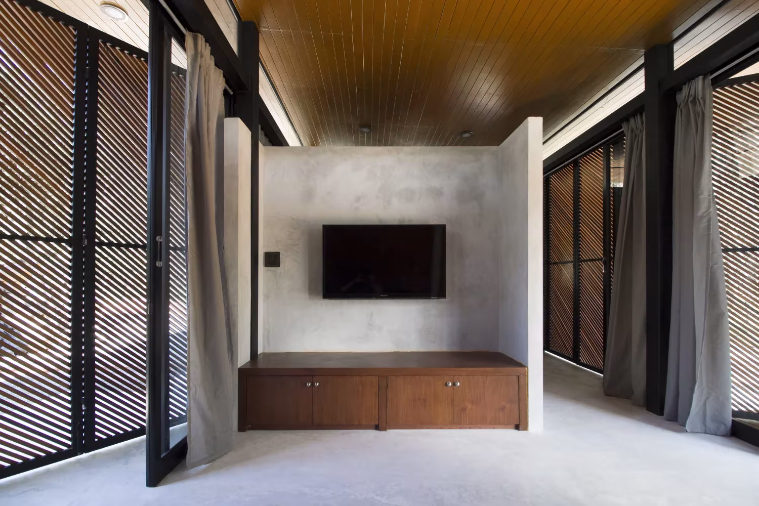 Estancia con mueble de madera y televisión entre celosías de lamas y cortinas en la Phatthalung House