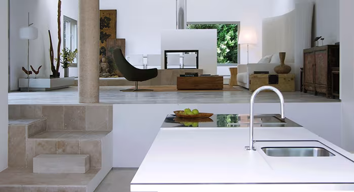 Cocina blanca abierta al salón en el proyecto C25, con isla de fregadero y butaca negra al fondo