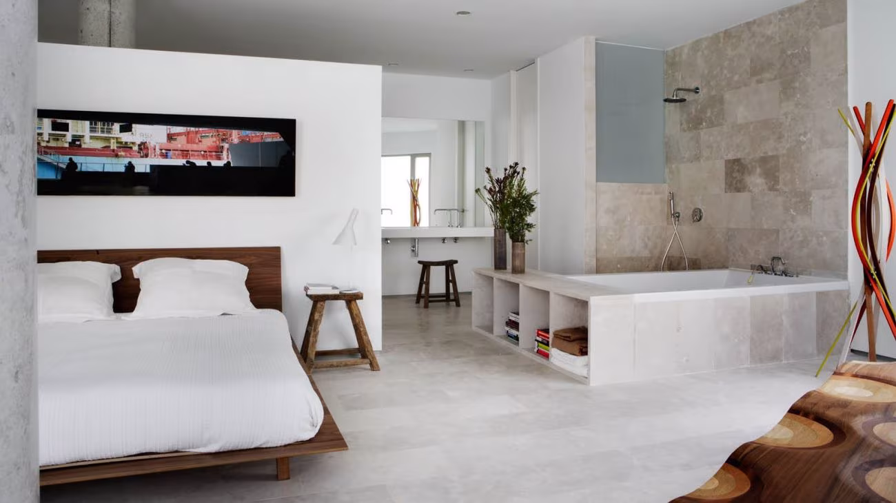 Dormitorio del loft Casa AA con cama de madera baja, paredes blancas y baño abierto con piedra al fondo