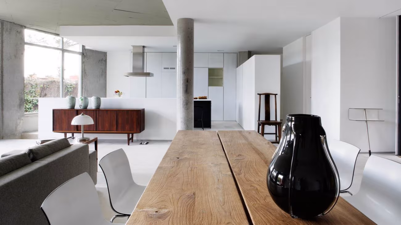 Salón diáfano del loft Casa AA con mesa de madera maciza, jarrón negro y cocina blanca al fondo