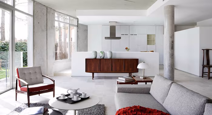 Salón tipo loft de la Casa AA con pilares de hormigón, sofá gris, aparador de madera y cocina blanca