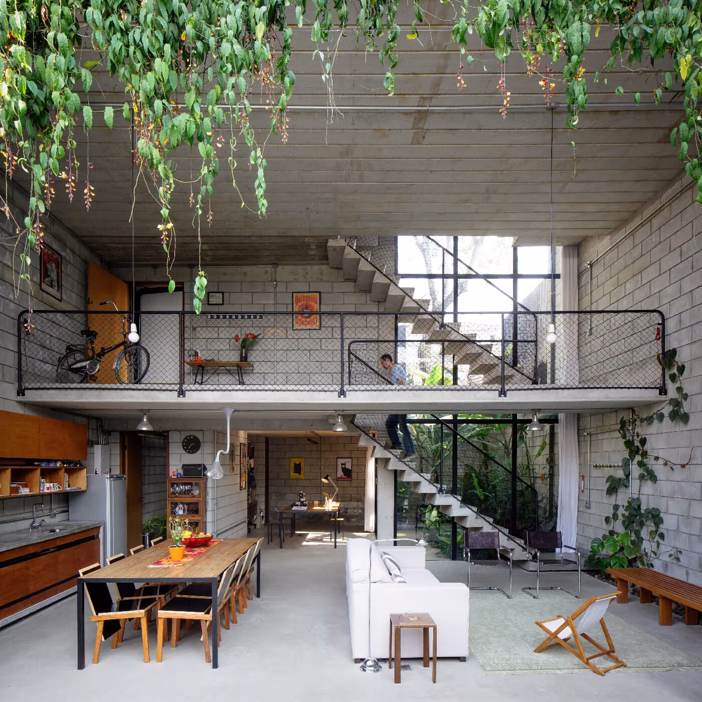 Salón de doble altura con plantas colgantes, entreplanta metálica y escalera de hormigón en la Casa Maracanã 81 de Terra e Tuma