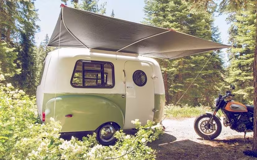 Caravana retro HC1 de Happier Camper con toldo desplegado en un pinar junto a una moto