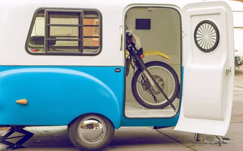 Caravana HC1 de Happier Camper azul y blanca con la puerta abierta y una moto guardada en su interior