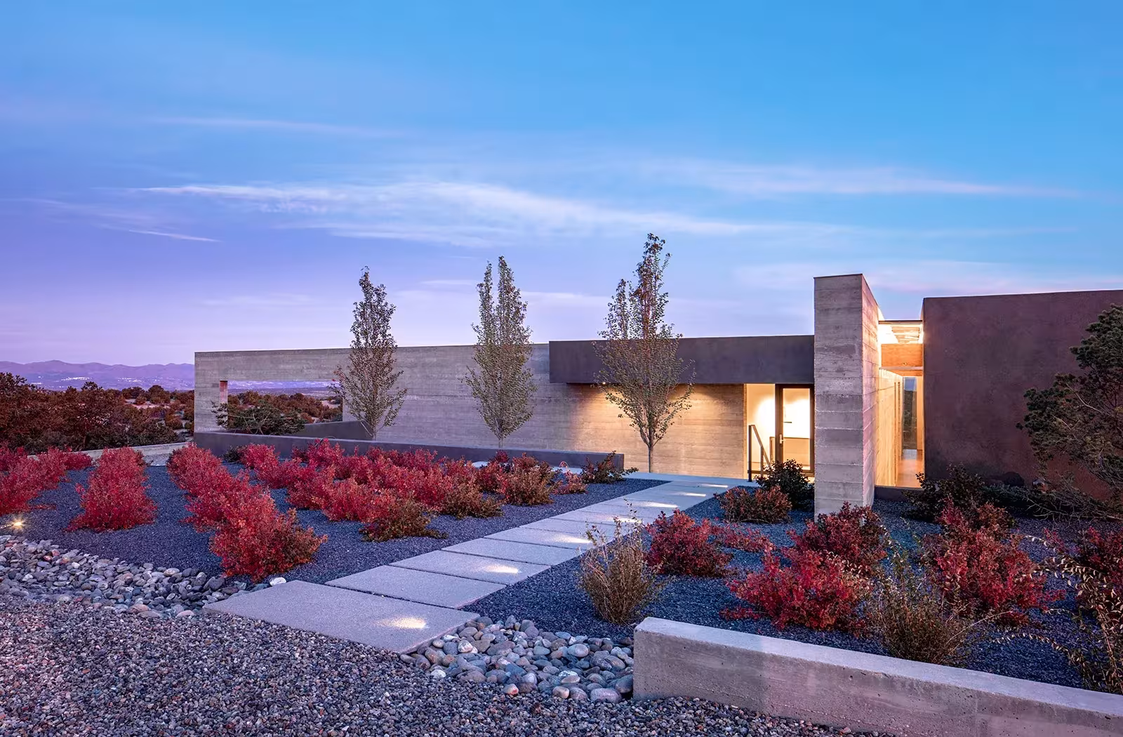 Sundial House al anochecer, con el muro de hormigón iluminado y un jardín de arbustos rojos en Nuevo México