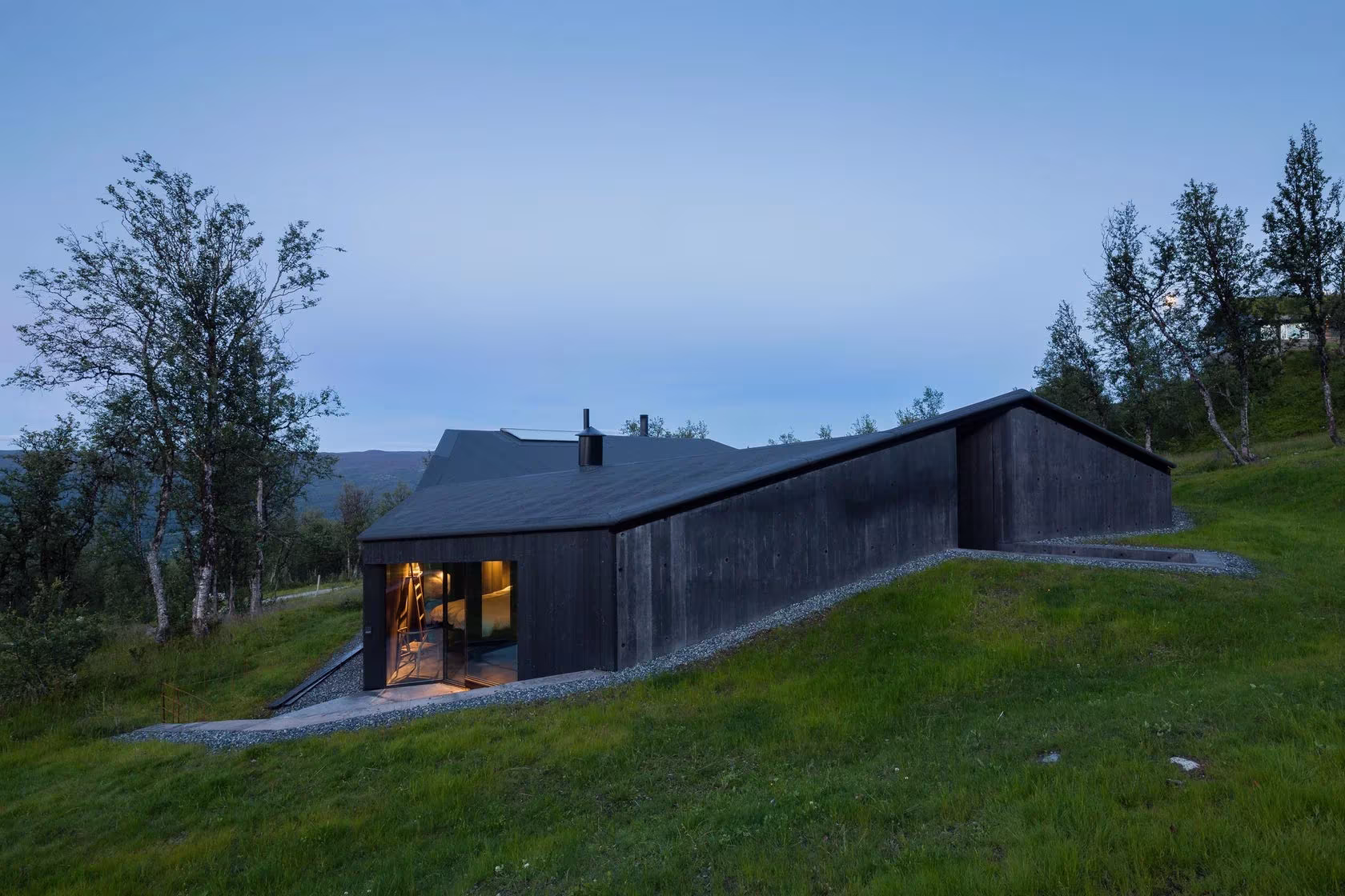 Cabaña Geilo hundida en la ladera cubierta de hierba al anochecer, con la fachada oscura iluminada