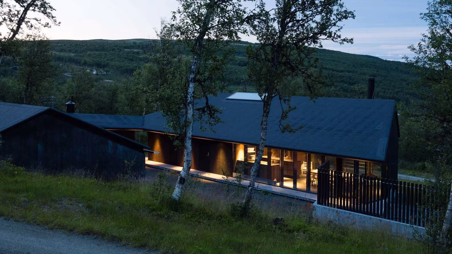 Cabaña Geilo con cubierta oscura a dos aguas y ventanales iluminados entre abedules, en las montañas noruegas