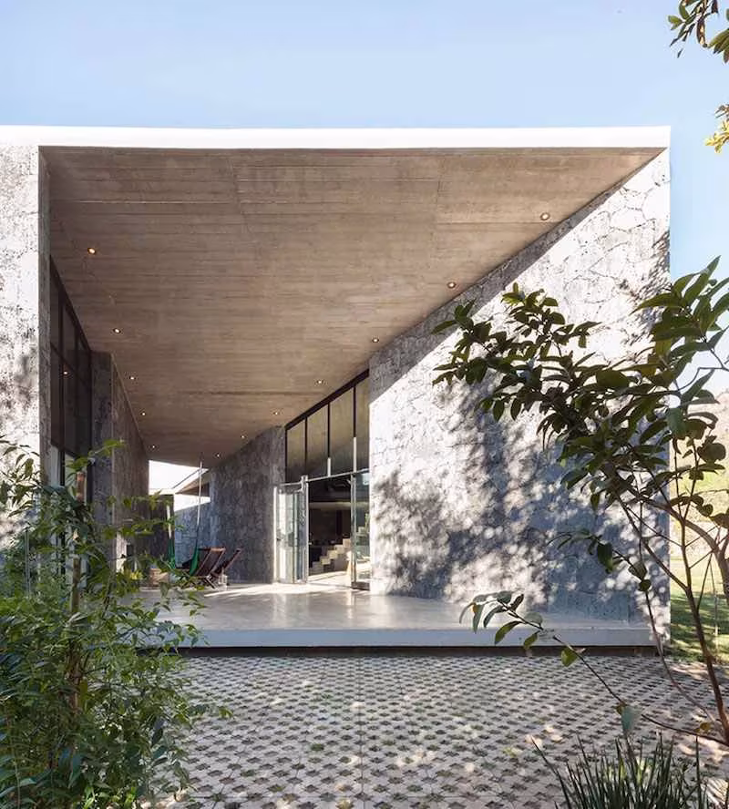 Fachada de piedra y hormigón de la Casa MA con la cubierta inclinada sobre un porche de adoquín ranurado