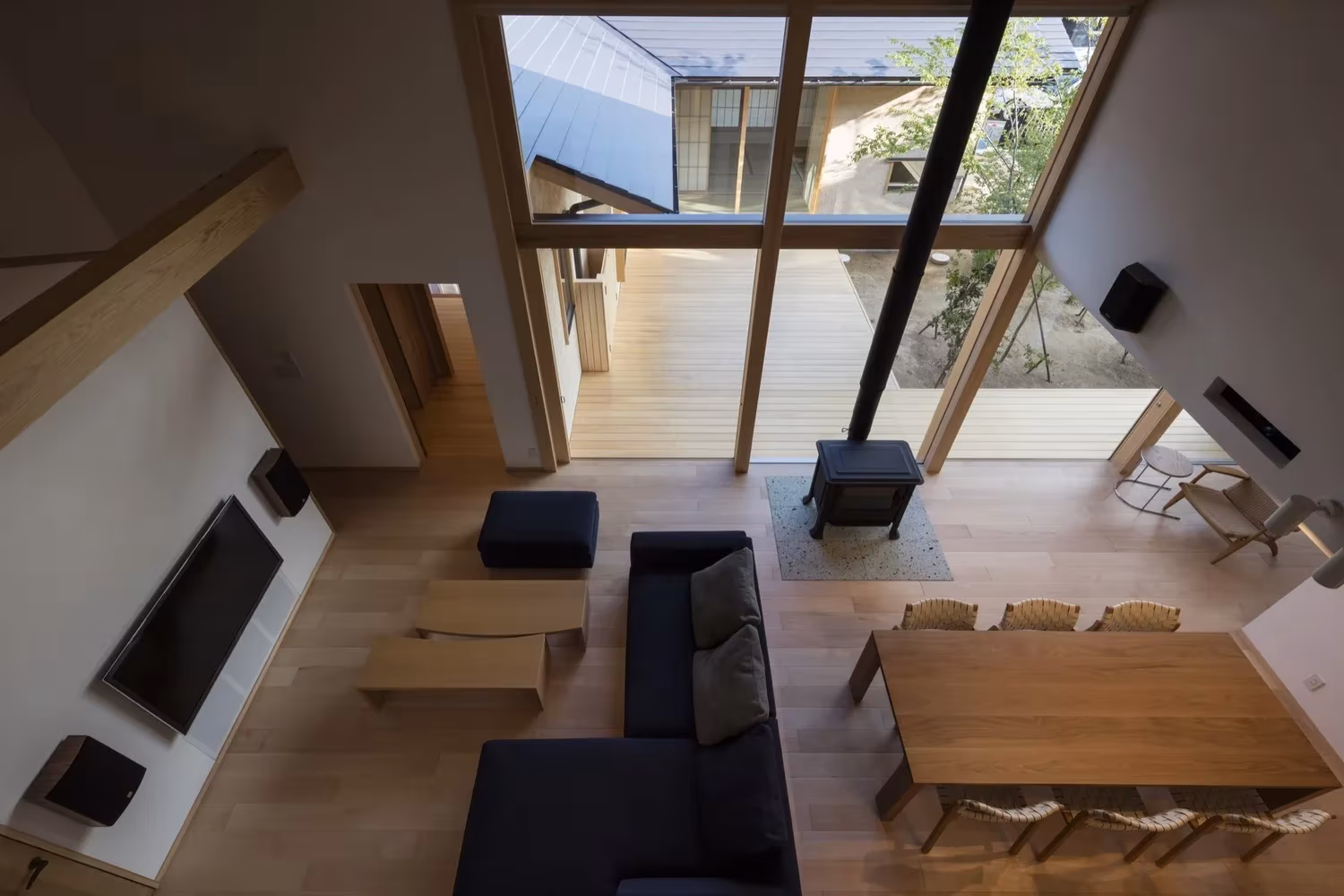 Vista cenital del salón con estufa, sofás y mesa de comedor bajo grandes ventanales en la Casa Hiiragi
