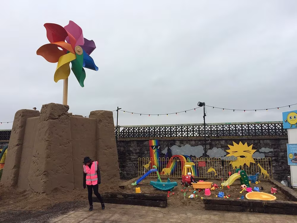 Castillo de arena con un molinillo gigante de colores y un arenero con juguetes en el parque Dismaland de Banksy