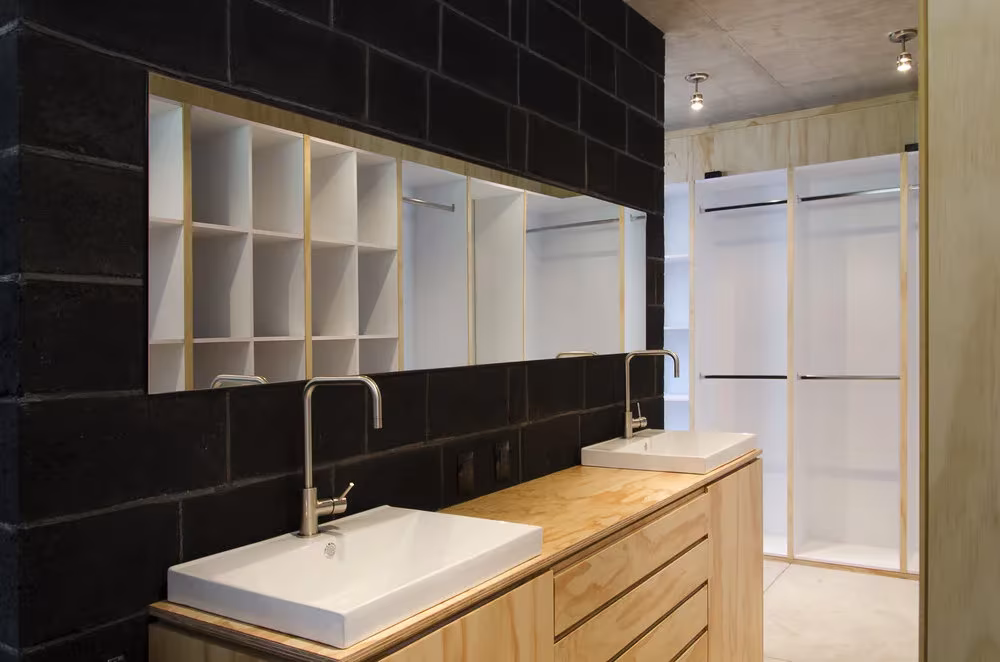 Baño con doble lavabo, muro de bloque oscuro y vestidor blanco con madera clara en la Casa BC de 3ARCH