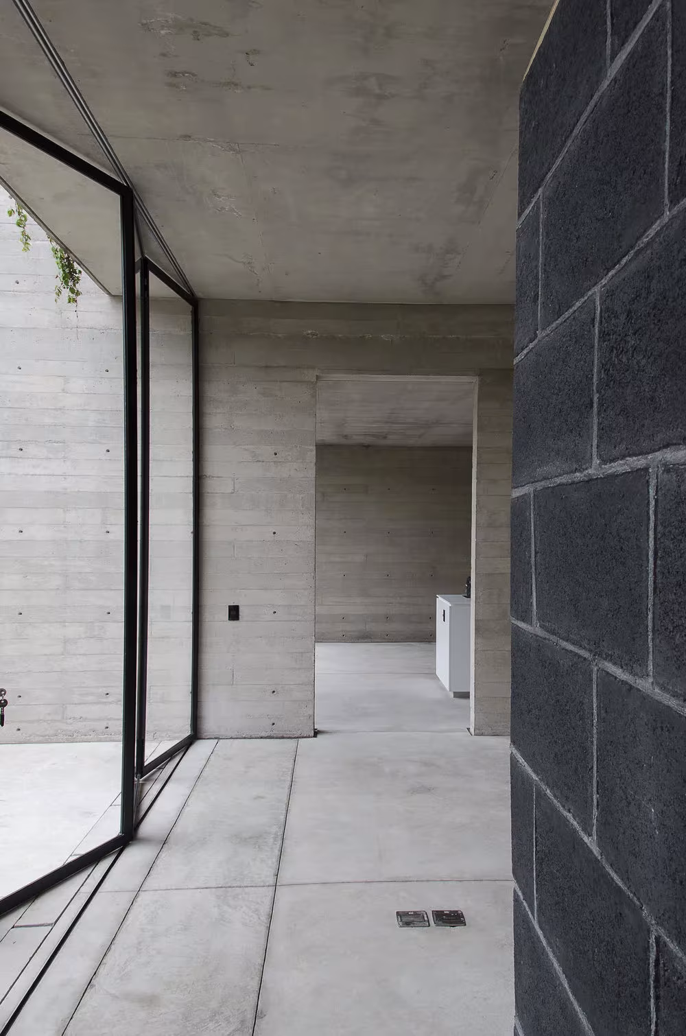 Vestíbulo de hormigón con muro de bloque negro y cristalera hacia el patio en la Casa BC de 3ARCH y Daniel Sampson
