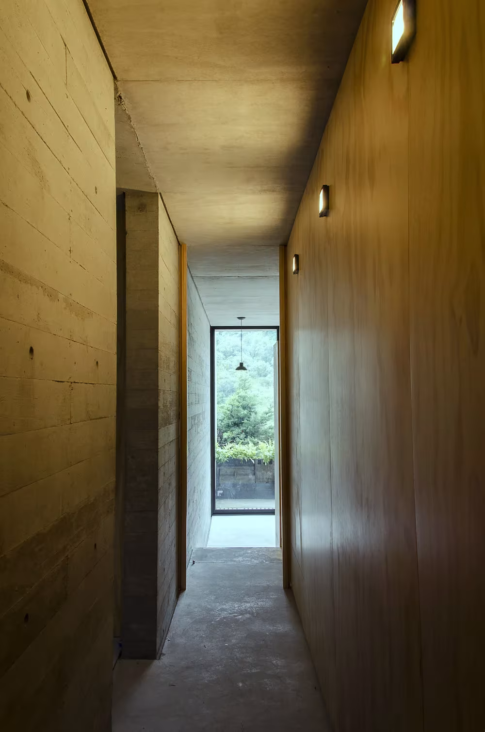 Pasillo estrecho de madera y hormigón con una ventana vertical al fondo en la Casa BC, Huixquilucan