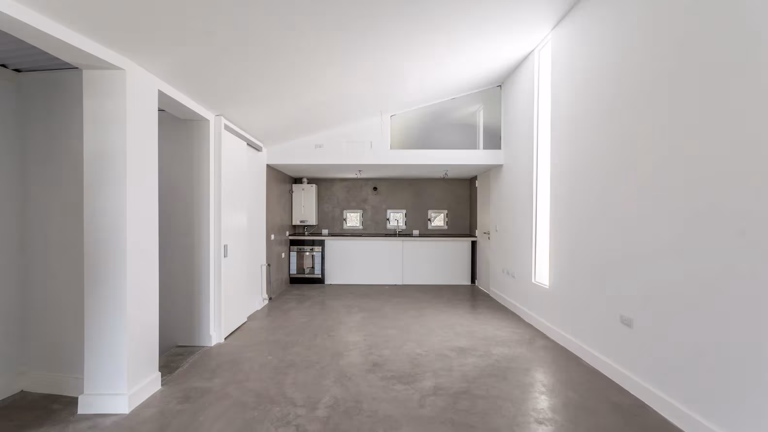 Interior blanco con suelo de cemento pulido, cocina al fondo y entreplanta bajo un techo inclinado en 5 Casas