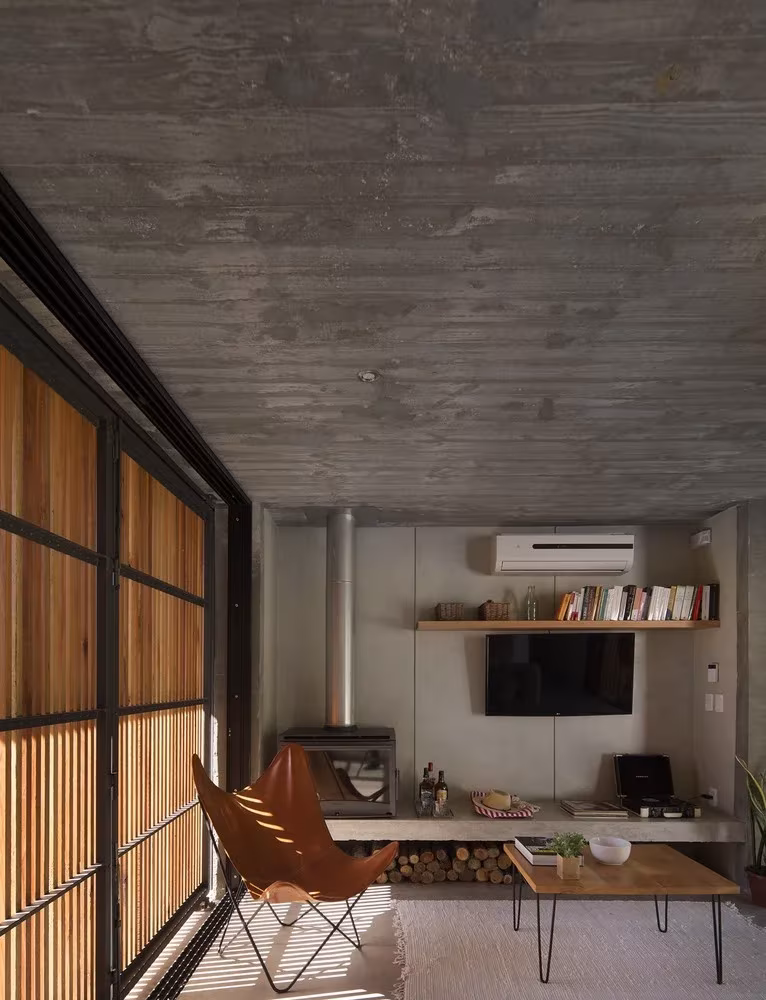 Sala con butaca de cuero, chimenea y ventanal de listones bajo el techo de hormigón de la Casa Marindia