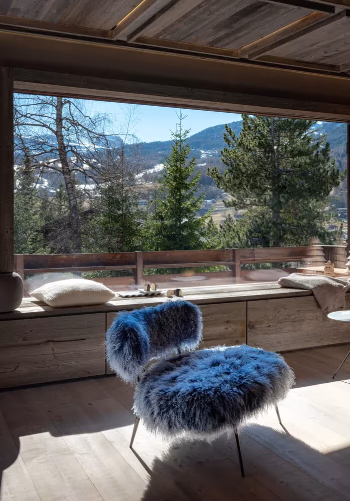 Casa Cortina de Outline Studio 74 con butaca de pelo azul ante un ventanal abierto a los bosques de los Dolomitas