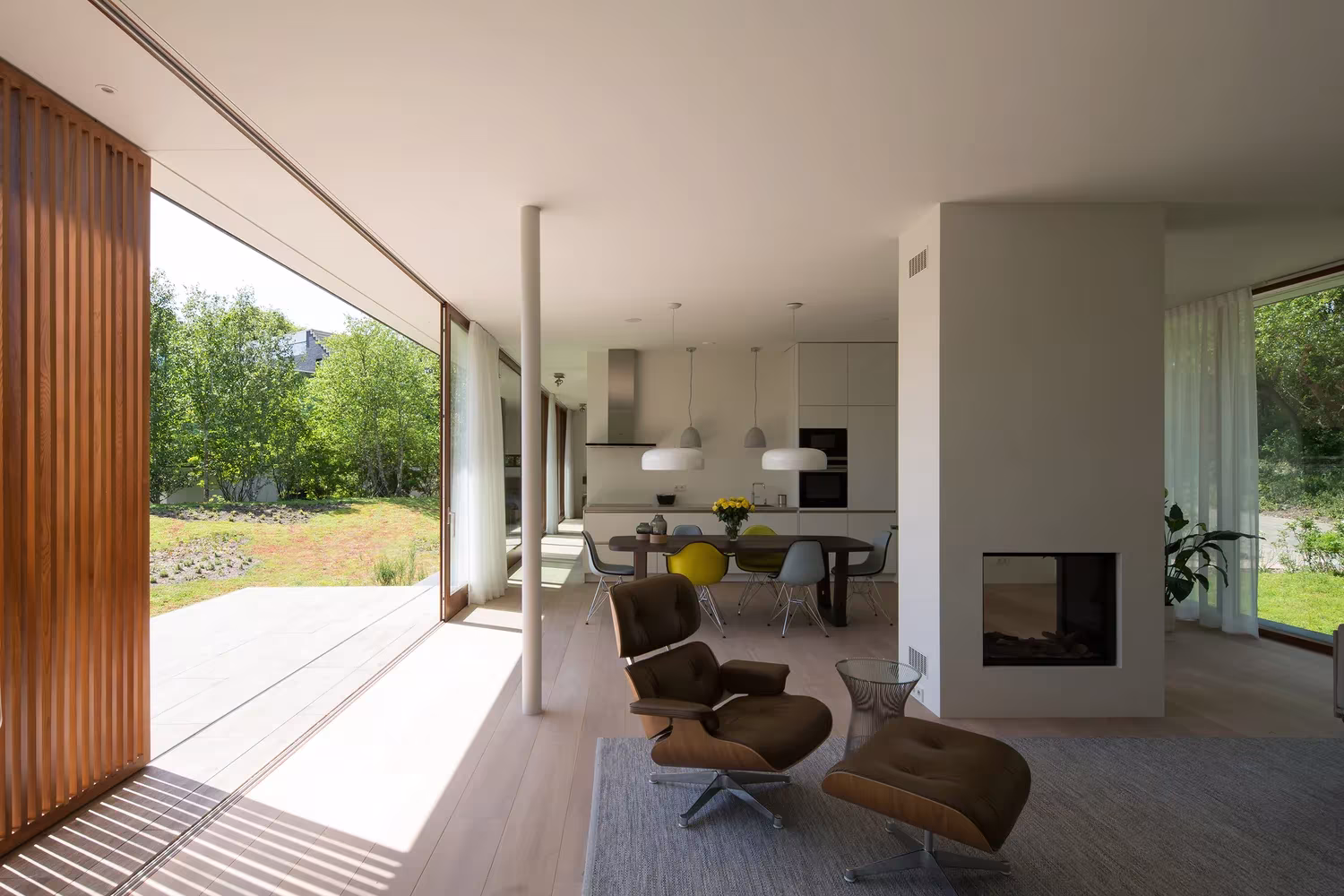 Salón con butacas de diseño, chimenea central y paneles de lamas de madera abiertos al jardín en la Villa H