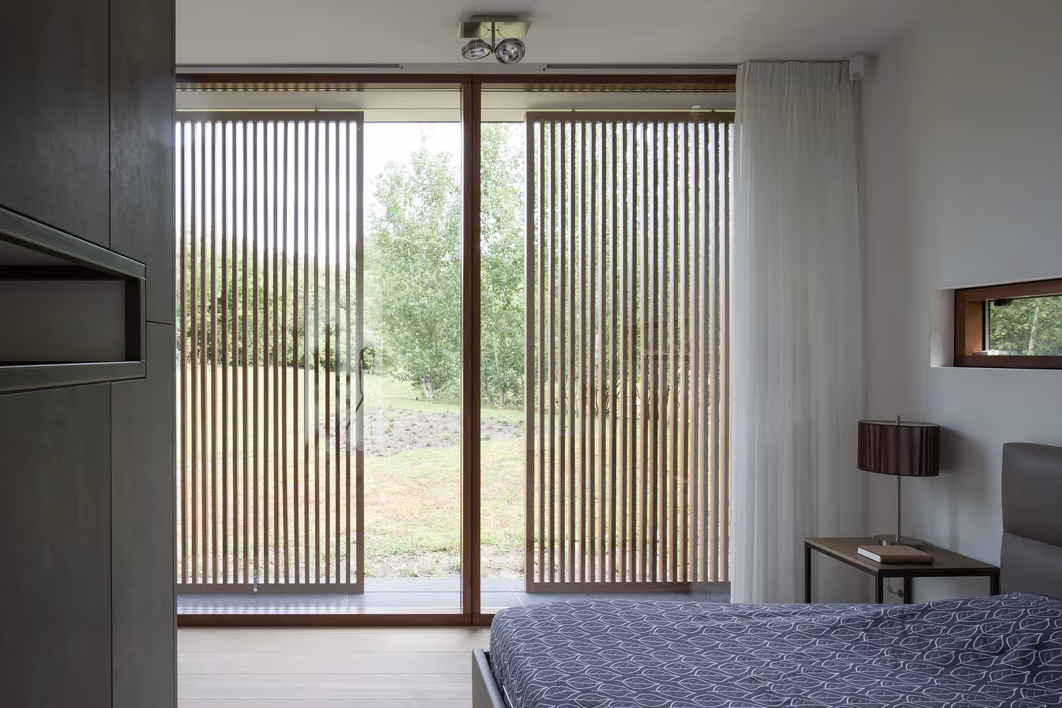 Dormitorio con puerta de vidrio y celosía de lamas de madera ante el prado en la Villa H de BERG + KLEIN