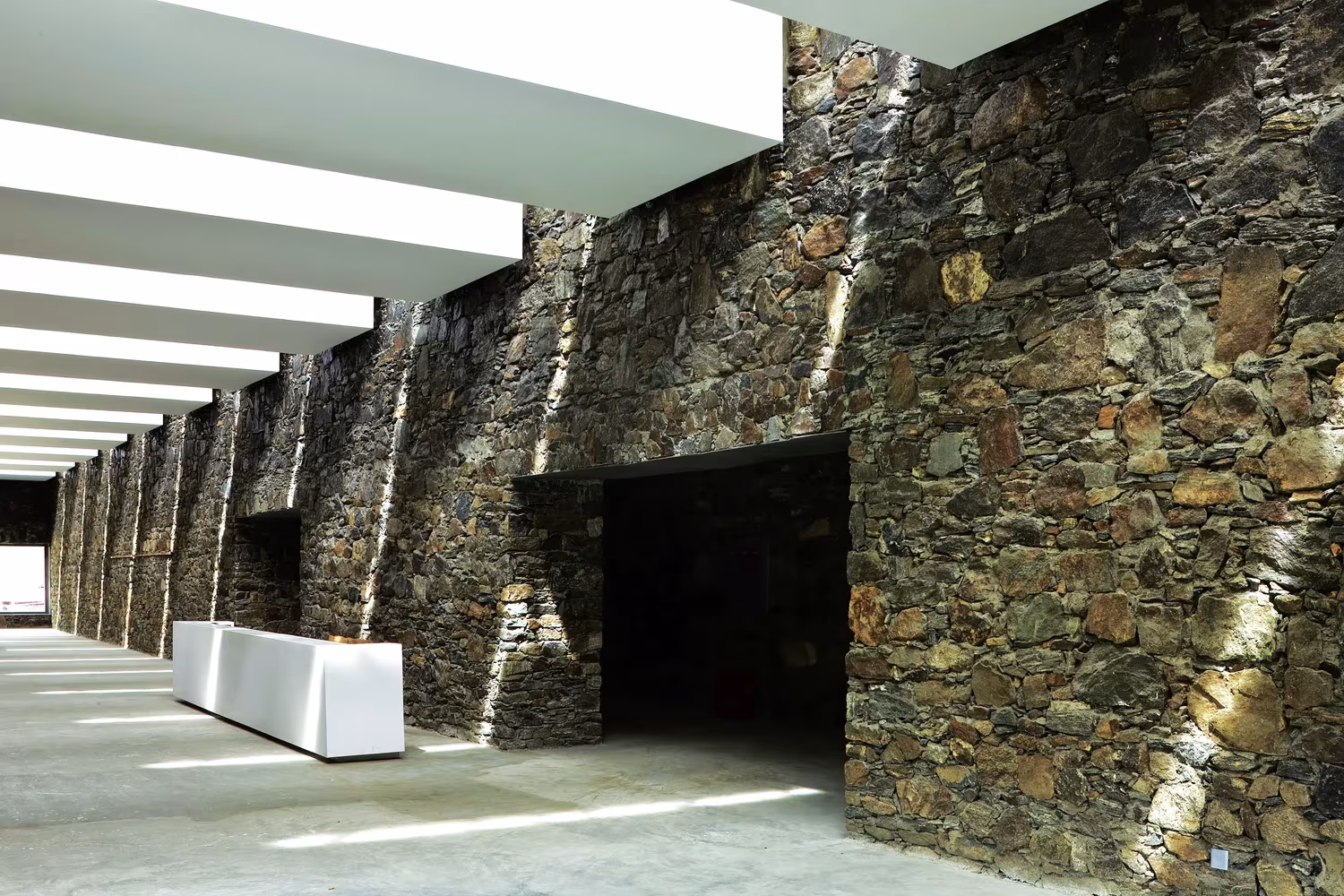 Vestíbulo del centro de visitantes de Namchabawa con muro de piedra, mostrador blanco y lamas de luz cenital