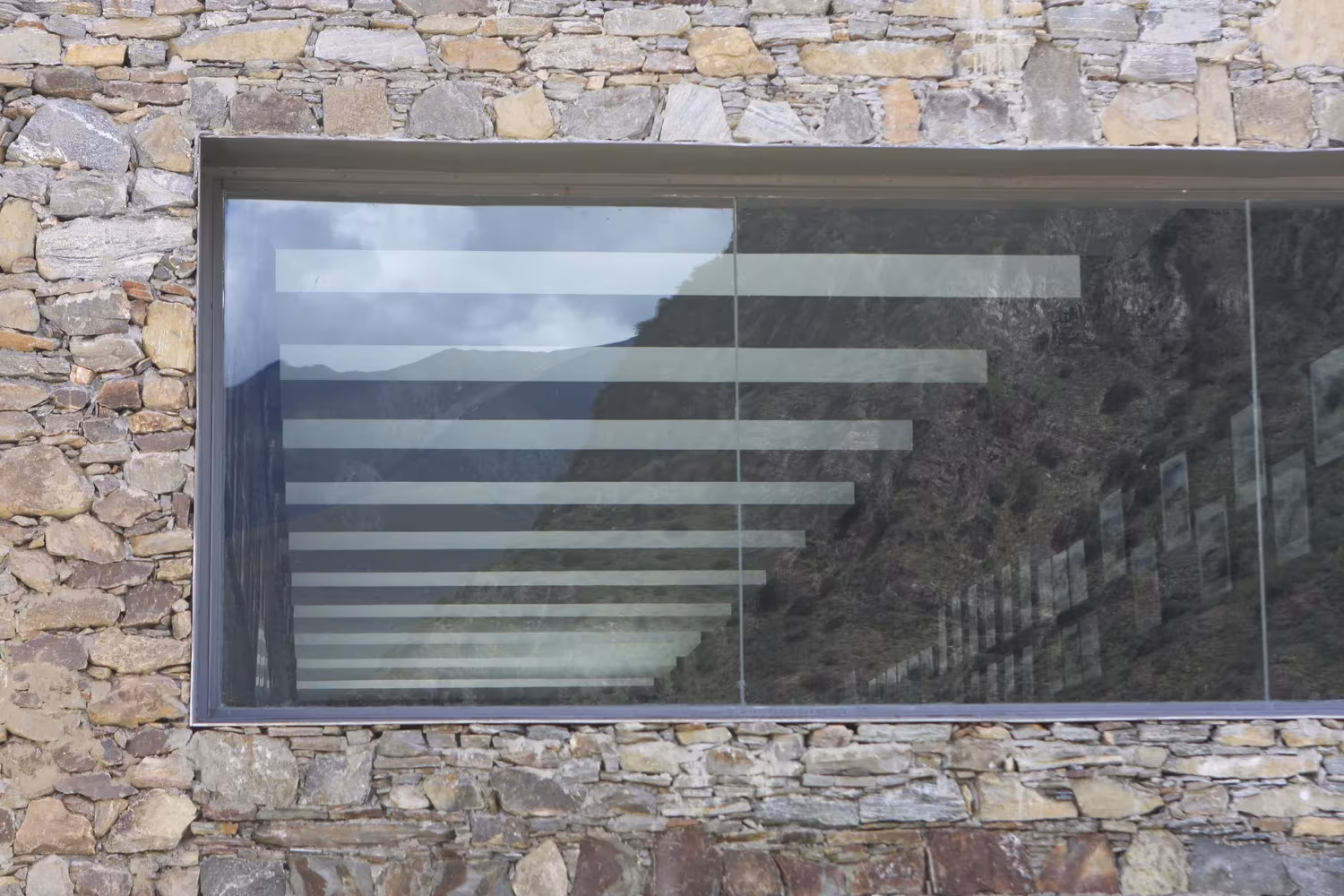 Ventana corrida enmarcada en muro de mampostería del centro de visitantes de Namchabawa que refleja las montañas