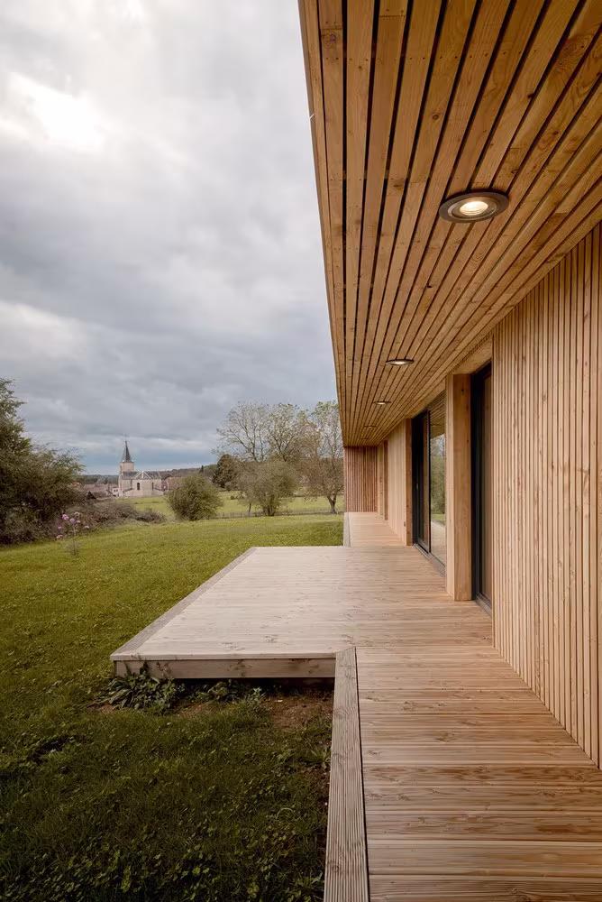 Porche de madera con tarima y techo de lamas sobre el jardín en la casa de Mathieu Noël, con la iglesia al fondo