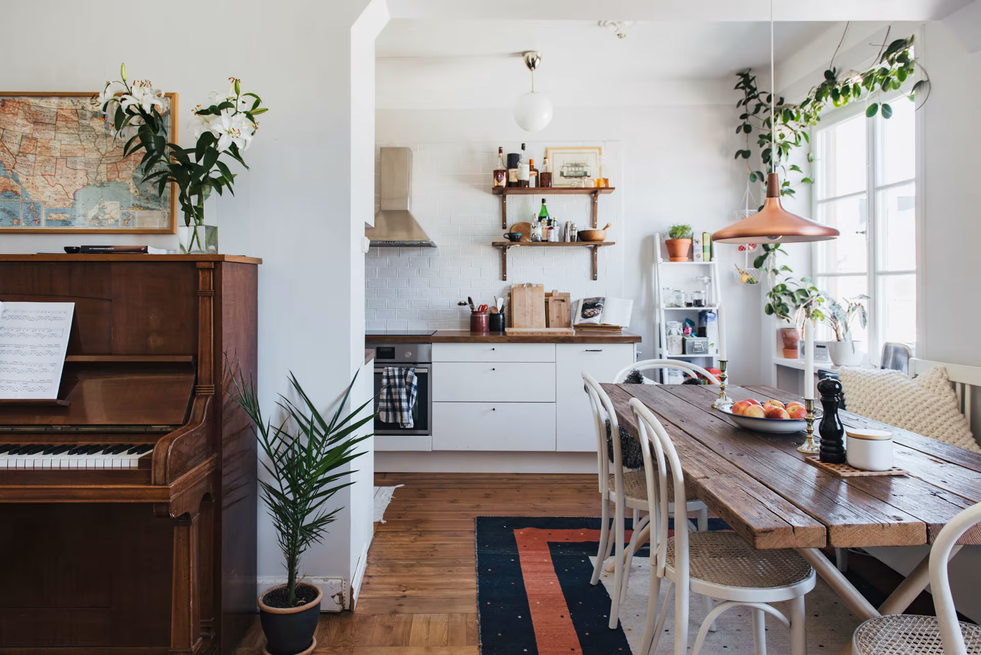 Cocina y comedor abiertos con mesa de madera, sillas blancas, estantes con plantas y un piano al fondo