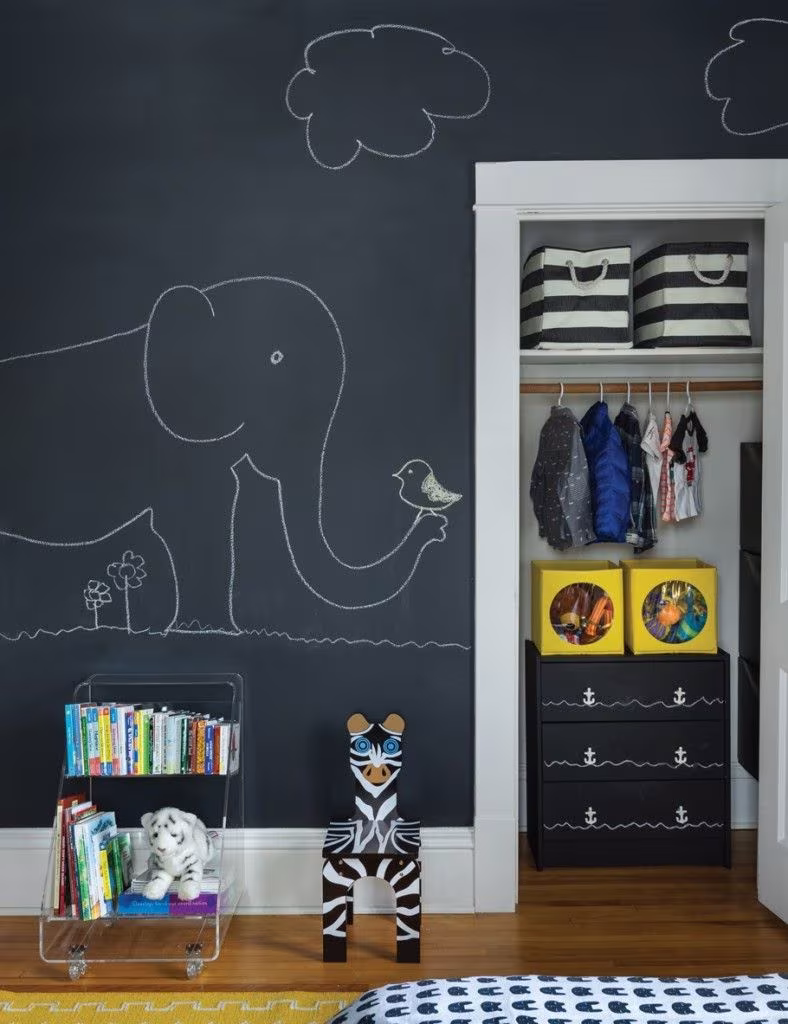 Pared de pizarra con dibujo de un elefante y una nube junto a un armario abierto en una habitación infantil