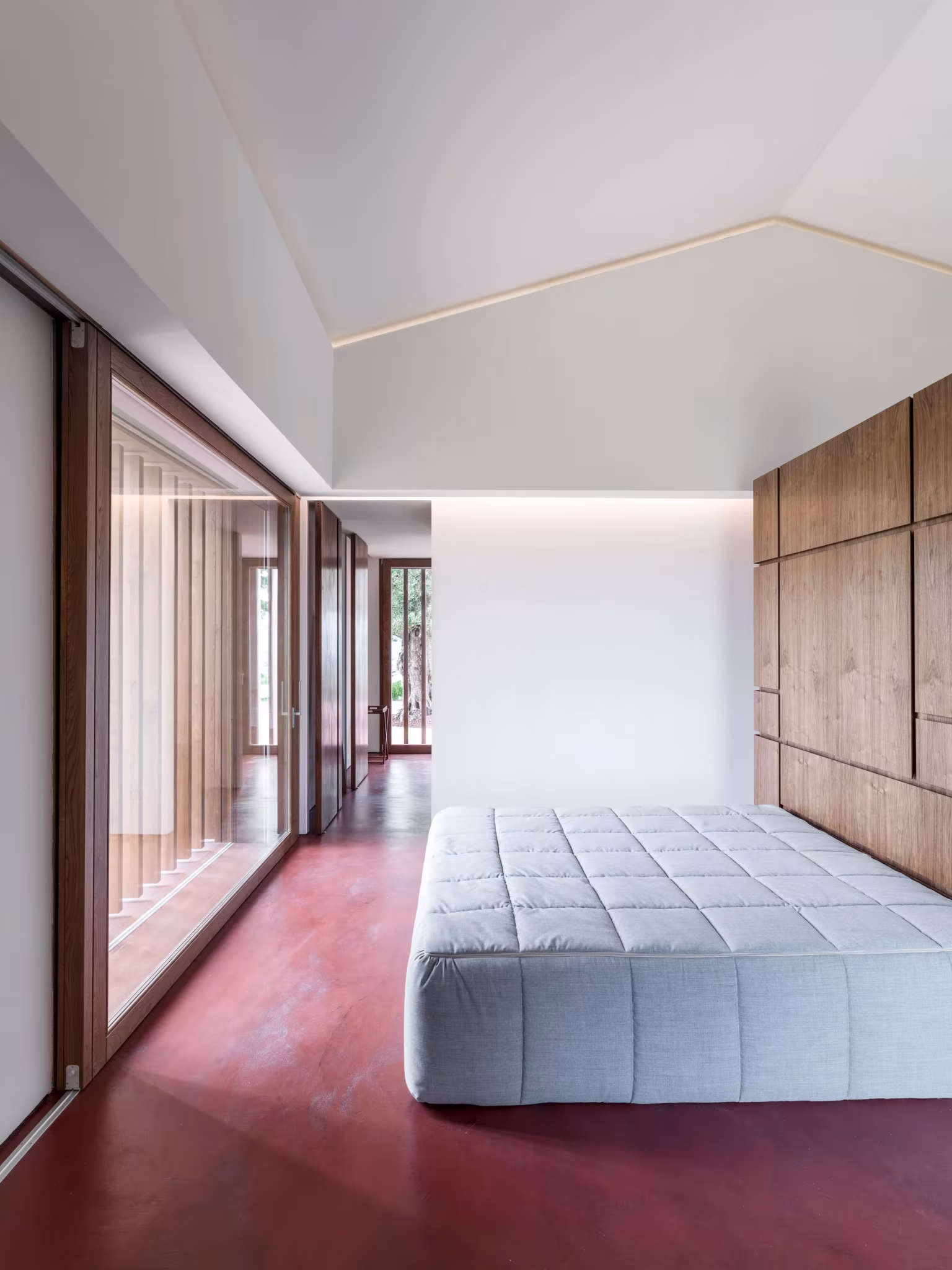 Dormitorio blanco con cama baja y gran ventanal corredizo sobre suelo rojo en la AP House de GGA Architects