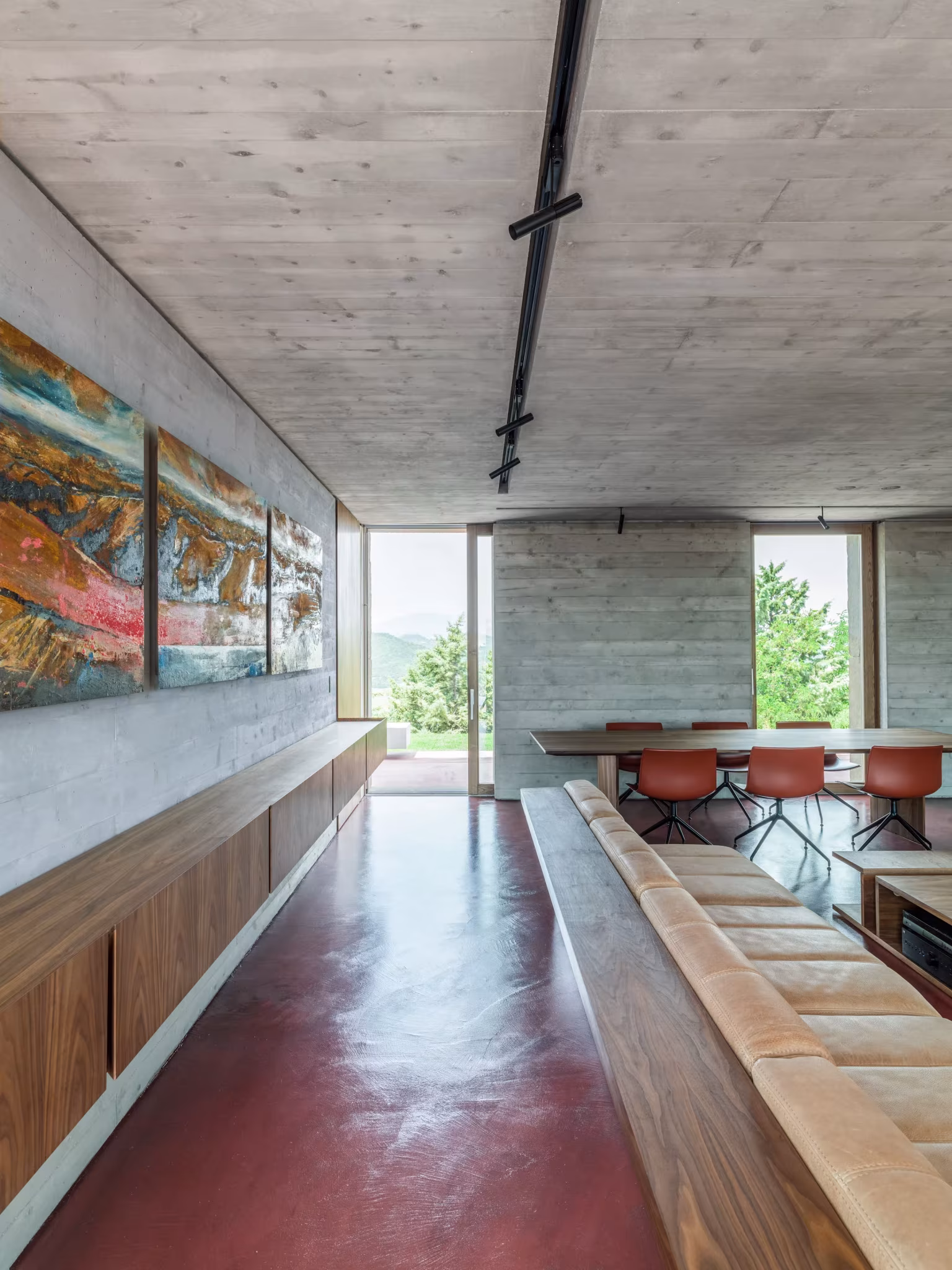 Galería interior de la AP House con techo de hormigón, suelo rojo y banco de madera hacia el comedor